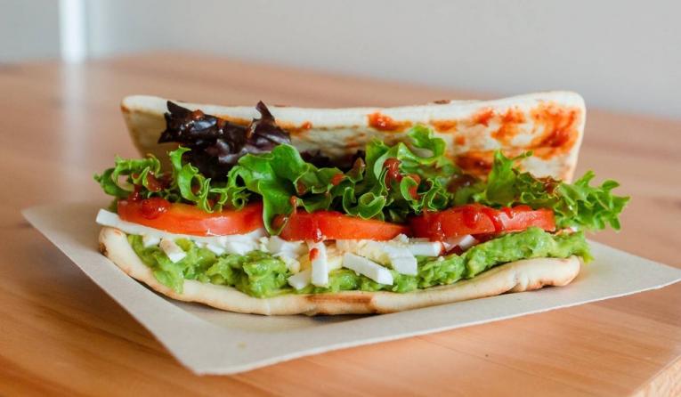 The Avo Egg Salad Flatbread Sandwich is a blend of avocado and hard-boiled eggs, evenly spread over a toasted panini flatbread with tomato slices, lettuce mix, and topped with a sriracha drizzle.